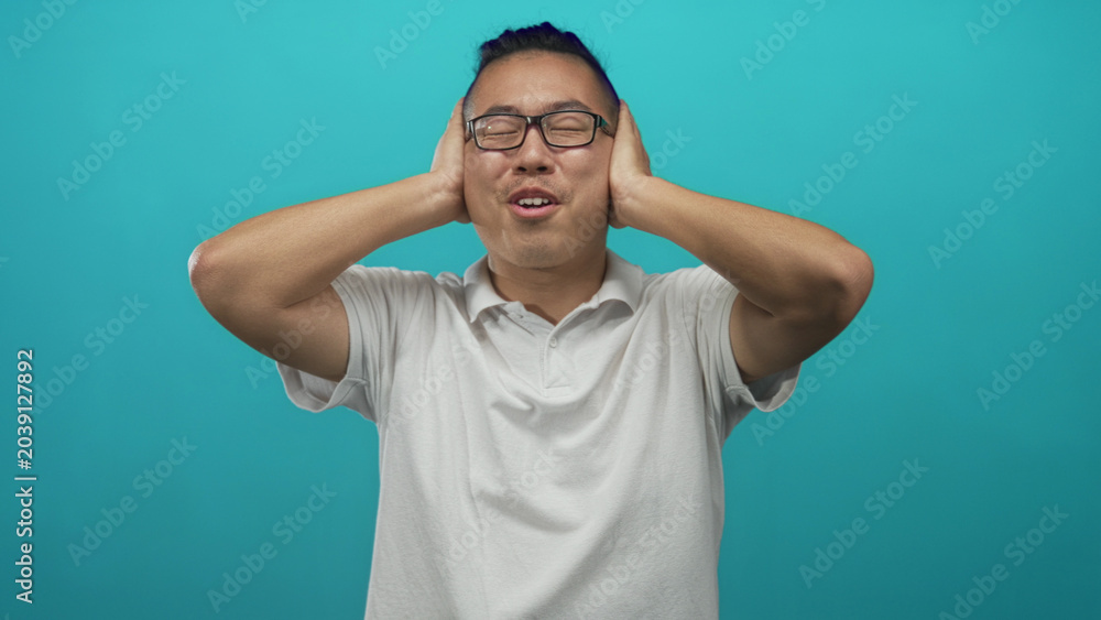 Fototapeta premium Man covering ears with hands and grimacing in a turquoise studio wearing white polo and glasses, hair tied in a topknot; discomfort.