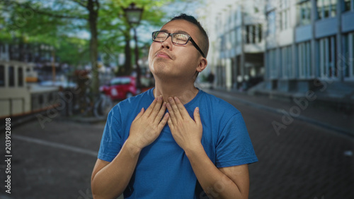 Foto Young chinese man in blue t shirt clutching neck and grimacing on a tree lined city street near canal and parked bicycles; discomfort
