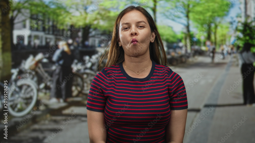 Fototapeta premium Woman makes fish face facing camera on city street with parked bicycles and leafy trees visible; playful mood.
