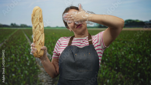 Tableau sur toile Woman wearing apron holds baguette aloft and covers eyes with gloved hand in green field; playful curiosity baking craft