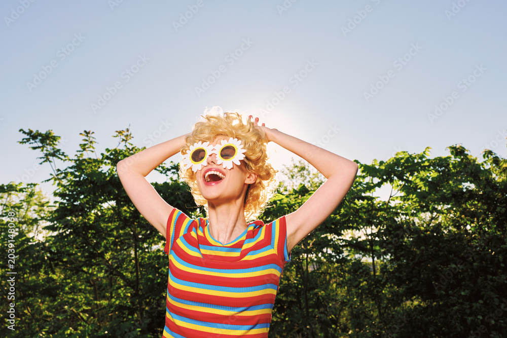Fototapeta premium Smiling woman with curly hair wearing flower glasses and a striped shirt in bright sunlight. Outdoor setting conveys playful summer vibes, carefree style, and bold color.
