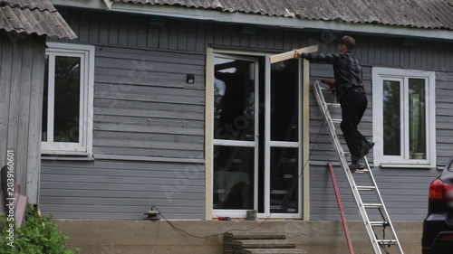 A man is renovating a wooden house, drilling a hole in the frame of a glass door outside the house