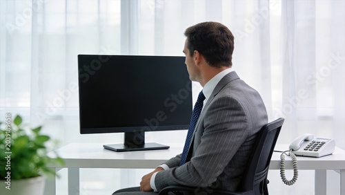 Businessman waiting at desk looking at blank computer monitor screen, side view of professional employee in suit sitting in office chair, empty display ready for content or
