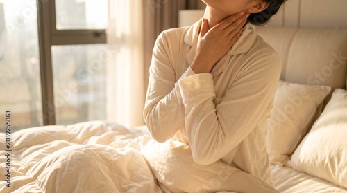 Sick woman touching neck feeling throat pain while sitting on bed at home