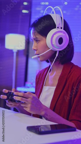 Excited young Asian girl playing video game with gaming headset and controller at home, esports online concept.