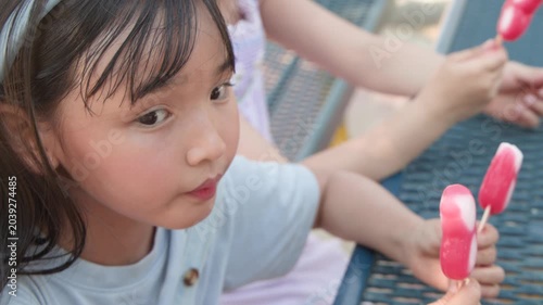 Two Young Asian Girls Enjoying Popsicles in a Sunny Outdoor Park