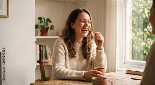 Happy woman in knitted sweater laughing at a table with coffee, inside a cozy home.