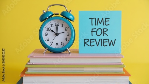 Time For Review note and blue alarm clock on a stack of books over a yellow background