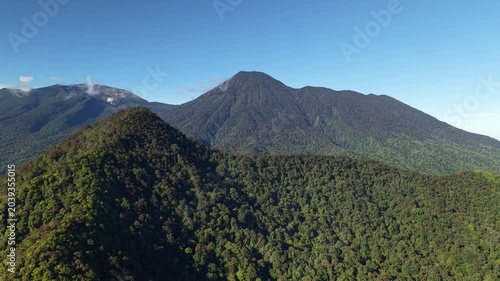 A reverse-motion aerial drone shot revealing a lush tropical forest and a landscape of layered mountains beneath a clear blue sky.