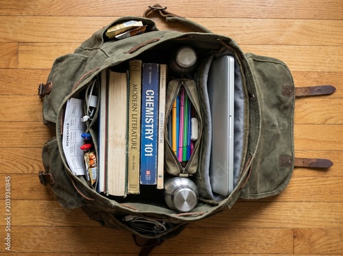 An open olive green canvas backpack neatly packed with textbooks, a silver laptop, and school supplies, resting on a wooden floor.