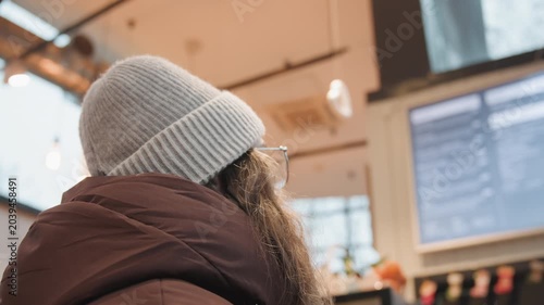 Woman facing cafe menu screens, indoor counter lights and menu display, knit beanie and padded jacket visible from back, quiet morning ordering moment, candid profile suggesting choice and warmth.
