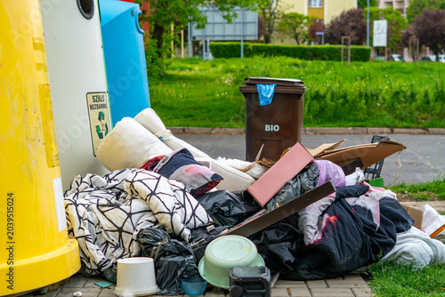 Bilde på lerret A pile of bulky waste, old textiles, cardboard boxes, and plastic trash bags abandoned on a pavement next to color-coded recycling bins, with a brown bio bin and green park in the background