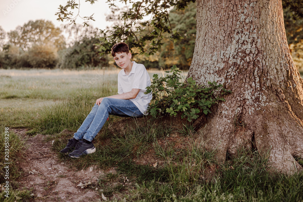 Obraz premium Teenage boy sitting beside large tree in peaceful meadow portrait
