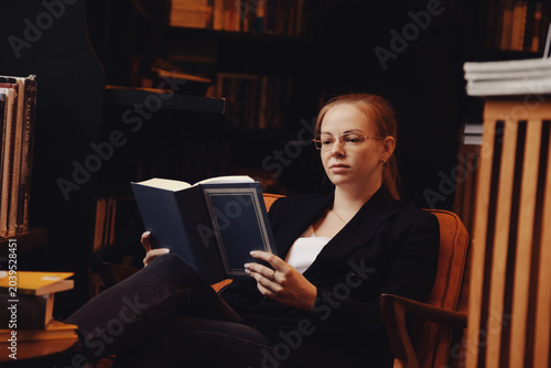 Фотография An adult woman sits comfortably in a library chair, deeply focused on a book
