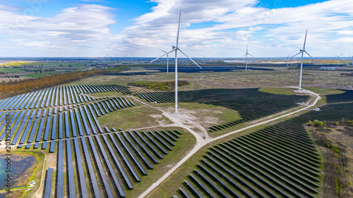 Fotografie Sunny, panoramic aerial drone view of a hybrid renewable energy farm featuring rows of blue solar panels and modern white wind turbines against green fields, a blue sky, and scattered clouds