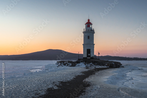 Bild auf Leinwand Lighthouse on frozen shoreline at sunset with snow-covered ground and distant mo