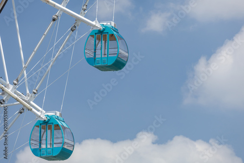Foto Two fully enclosed gondola cabins hang from a Ferris wheel's white support beams, set against soft fluffy clouds on a clear blue sky, embodying concepts of leisure as well as aspiration and freedom