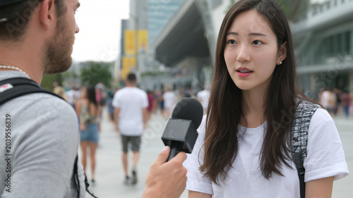 Lovely charming girl giving interview answering to journalist in microphone public asking broadcast outdoors. Multinational people being interviewed on the street by the media.
