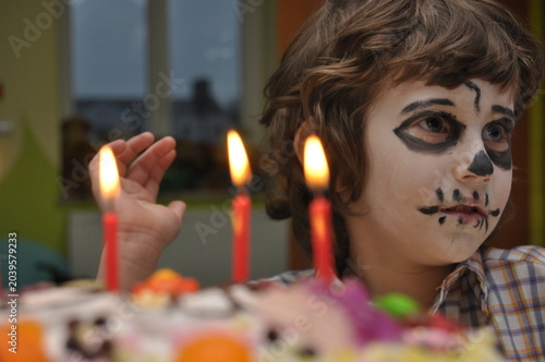 Child celebrating birthday with face painted like a skeleton near a colorful cake and lit candles in a festive setting