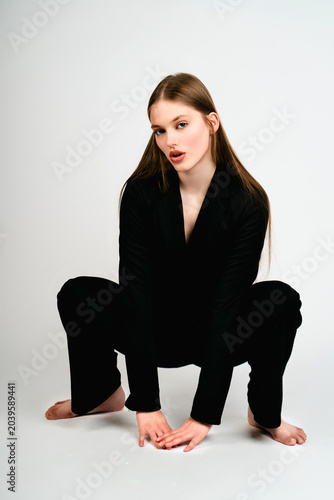 Bilde på lerret Full-length studio portrait of an elegant young woman crouching in a black pantsuit on a solid white background
