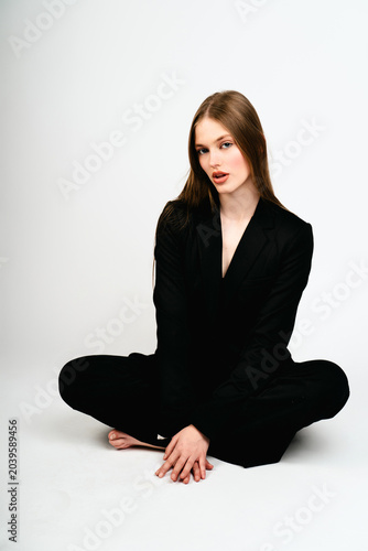 Quadro su tela Young female model wearing a black formal pantsuit sitting cross-legged on a clean white background in a studio setting