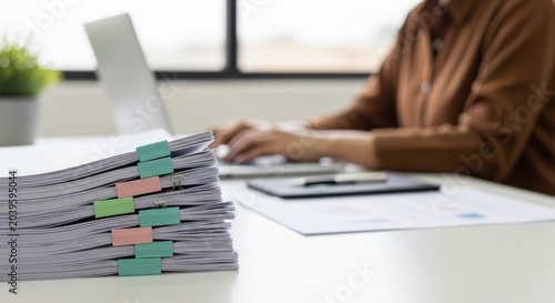 Stack of documents in binders on desk with person working on laptop in blurred background at bright office for administrative work and organization concept