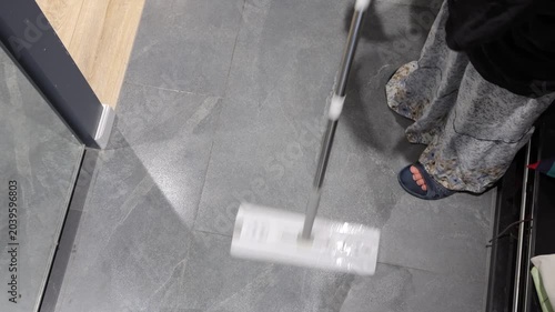 A teenager guides a flat mop over gray tile near a wooden floor threshold during entryway cleaning.