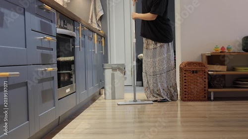 A teenager cleans the kitchen floor with a flat mop beside lower cabinets and a gray bucket.