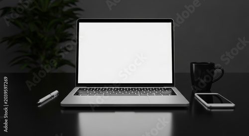 Modern silver laptop computer with blank white screen display for website mockup design standing on dark desk with smartphone and coffee mug in office workspace