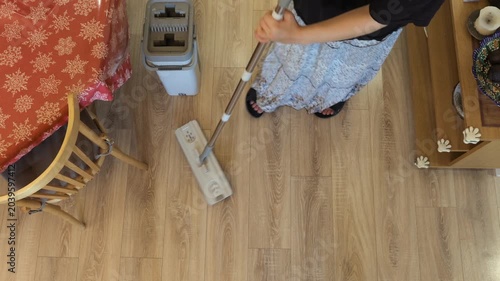 A flat mop moves across a light wooden floor beside a gray bucket in an indoor cleaning task.