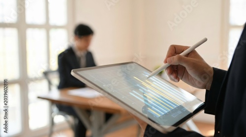 Business analyst using digital tablet with electronic pen to analyze financial data charts in modern office workspace while colleague working in background bokeh