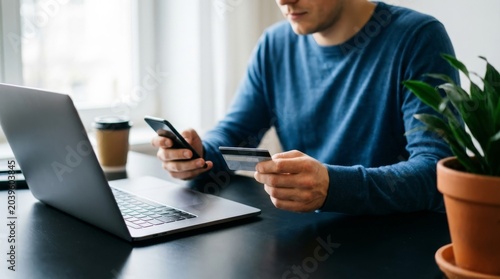 Young man using credit card for online shopping payment while holding smartphone and sitting at desk with laptop computer at home during day secure transaction