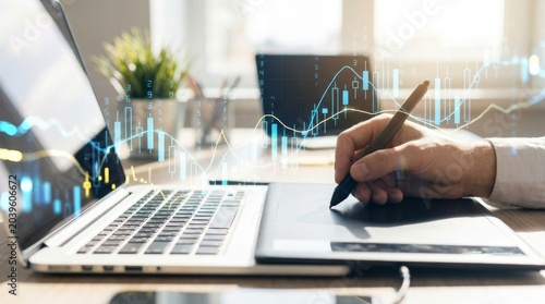 Male hands using digital graphics tablet and stylus pen with laptop for financial data analysis showing holographic growth charts and stock market trading statistics