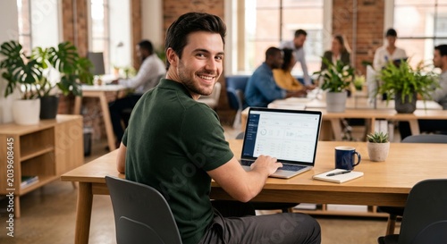 Happy young male entrepreneur working on laptop looking back at camera in modern open plan office with diverse team