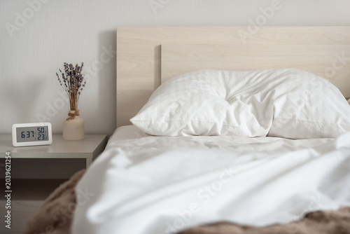 Minimalist bedroom interior with white bedding, bedside table and digital thermometer showing temperature and humidity. Cozy home environment and healthy sleep concept.