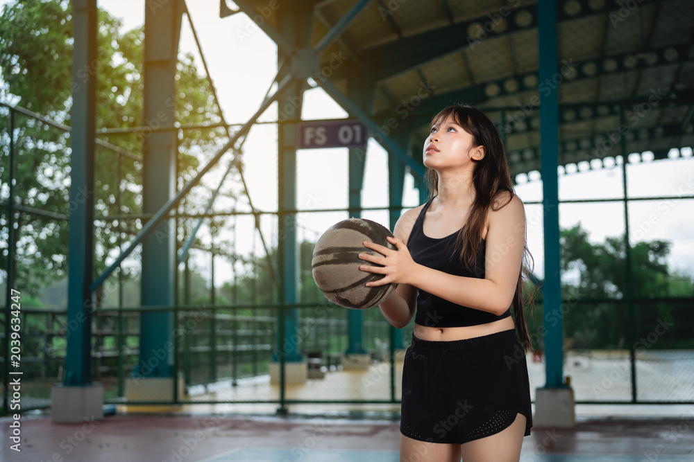 Naklejka premium Young woman in athletic wear holding basketball on outdoor sports court during golden hour, preparing for exercise or game, with a covered roof structure and greenery in background