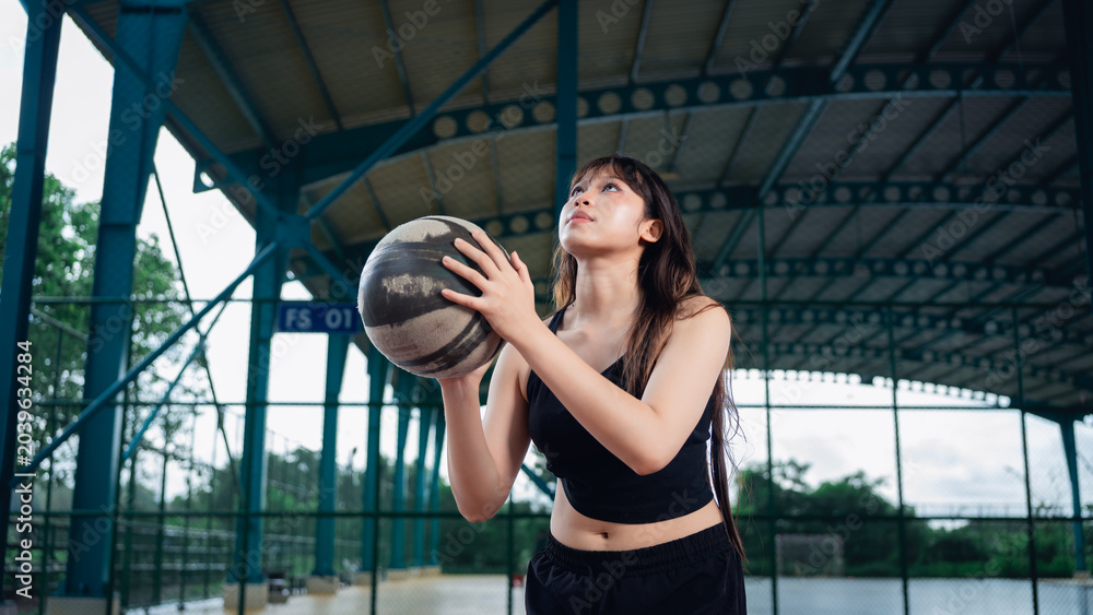 Naklejka premium Young woman in athletic wear holding basketball on outdoor sports court during golden hour, preparing for exercise or game, with a covered roof structure and greenery in background
