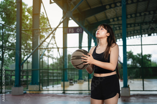 Young woman in athletic wear holding basketball on outdoor sports court during golden hour, preparing for exercise or game, with a covered roof structure and greenery in background