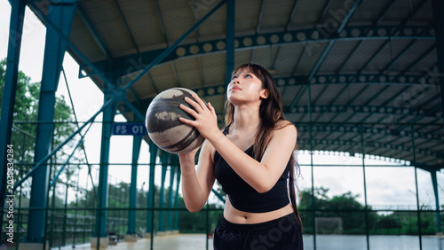 Young woman in athletic wear holding basketball on outdoor sports court during golden hour, preparing for exercise or game, with a covered roof structure and greenery in background