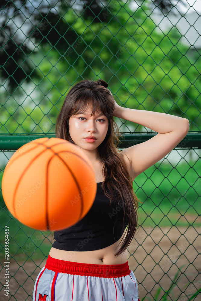 Naklejka premium Young woman with long hair and casual attire posing on a basketball court near a chain-link fence with a orange basketball in front of her and lush green trees background