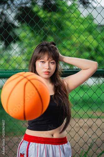 Young woman with long hair and casual attire posing on a basketball court near a chain-link fence with a orange basketball in front of her and lush green trees background