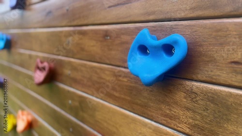 A close-up shot of a blue heart-shaped climbing hold on a wooden wall, with other colorful holds blurred in the background.