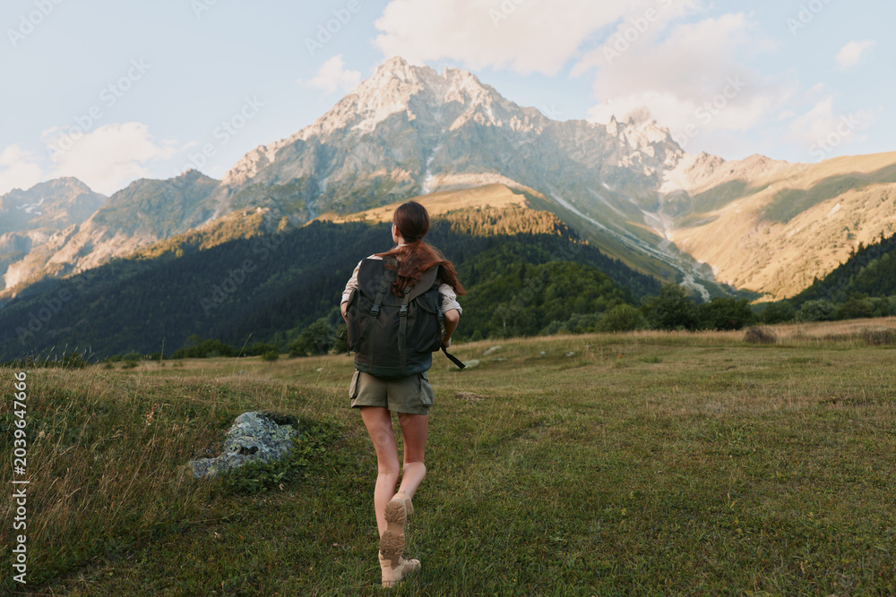 Naklejka premium A young woman with a backpack walks across a grassy meadow toward distant rugged mountains, embodying adventure and freedom in a vast natural landscape under clear skies