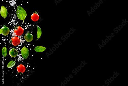 Fresh cherry tomatoes with green basil leaves and falling sea salt on black background with right copy space for vegan restaurant menu banner