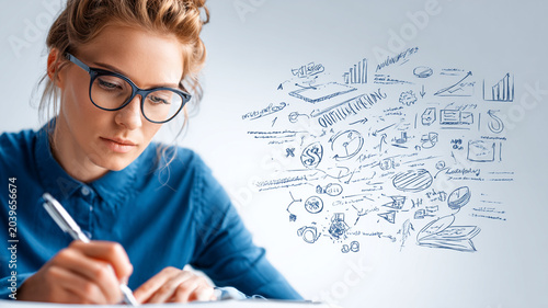 Focused woman in glasses writes notes with creative marketing strategy sketches on blue background, symbolizing innovation and planning