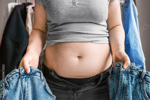 Fotografija Overweight woman holding small jeans in front of her bare belly, experiencing clothing annoyance and wardrobe problems due to weight gain