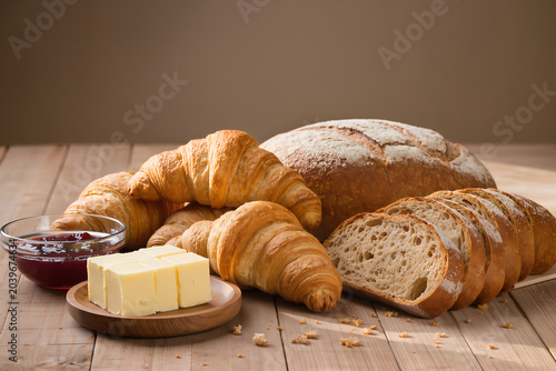 Freshly baked artisanal bread assortment with flaky croissants, sliced sourdough loaf, creamy butter cubes, and sweet berry jam on a rustic wooden table top background for breakfast food styling conce