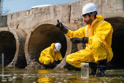 Two ecologist technicians in yellow hazmat suits inspect industrial wastewater pipes. One kneels looking into a concrete stormwater pipe, another holds a sample container