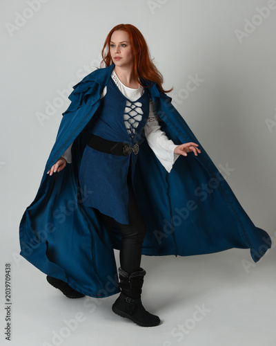 full length portrait redhead female model wearing medieval fantasy warrior historical costume with blue hood cloak. walking away with magical spell arm gesture, silhouette figure on studio background.