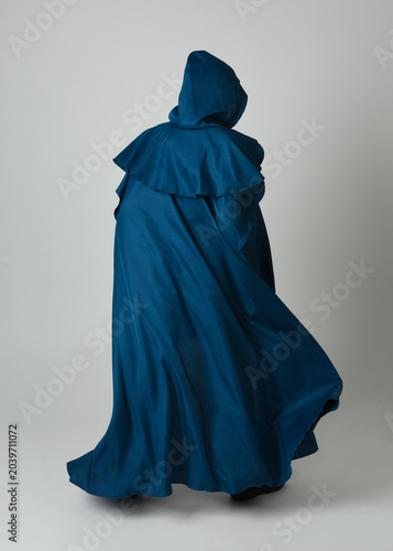 full length portrait of model wearing medieval fantasy warrior historical costume with blue hooded cloak. silhouette figure,  walking away in back view on studio background.